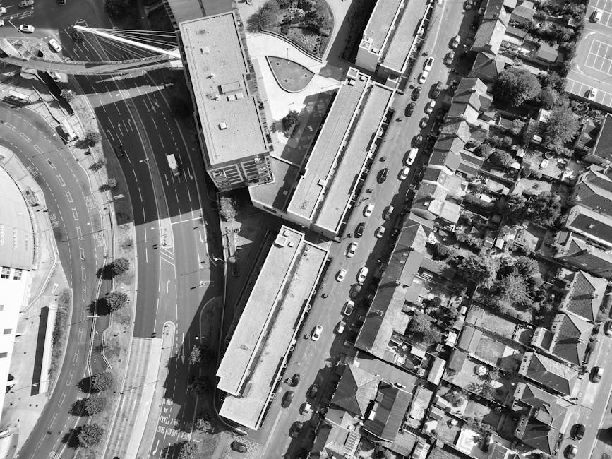 Aerial black and white photograph showing a narrow residential street in Whitechapel, lined with terraced houses on one side and modern buildings on the other. Several cars are parked along the curb, and a row of large cardboard boxes, wrapped furniture, and packing materials are arranged on the pavement, ready for a home relocation. A moving van is parked at the side of the street, with a few boxes and packing blankets nearby, indicating the loading process. The image captures the urban environment during daylight, with shadows cast by structures and trees, illustrating the logistical considerations involved in house removals and furniture transport. This scene reflects the planning and organization required for efficient packing and loading in busy Whitechapel locations, often managed by services like Man With a Van Whitechapel during moving days.