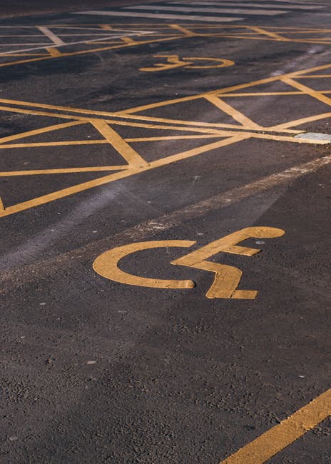 A close-up view of a parking lot surface featuring yellow painted accessibility and disabled parking space markings on dark asphalt. The symbols include the international wheelchair symbol and diagonal lines indicating reserved areas. In the background, additional yellow lines and markings designate adjacent parking zones. The surface appears slightly worn with visible patches of dirt and minor cracks. The image provides a clear view of the parking layout, suitable for illustrating parking access considerations for house removals and local logistics, as offered by Man With a Van Whitechapel, especially in relation to planning for home relocation and furniture transport in urban environments.