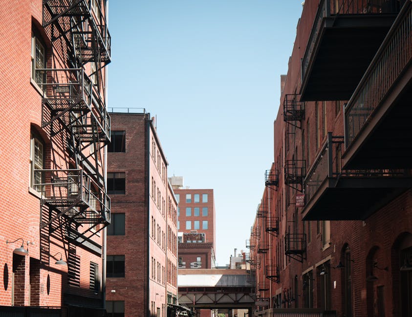 View of a narrow urban alleyway between red-brick residential buildings during daylight, with fire escape ladders and balconies made of black metal on both sides, casting shadows on the brick facades. A white van with a partially visible roof is parked at the pavement, with a person inside loading cardboard boxes and wrapped furniture using plastic and blankets. The boxes are stacked inside and near the vehicle, some on a trolley, as part of a home relocation process. The environment features clear blue sky overhead, natural lighting highlighting the textures of the brickwork and metalwork, and the scene captures a furniture transport and packing stage typical of house removals in the Whitechapel area, with Man With a Van Whitechapel providing professional removals service.
