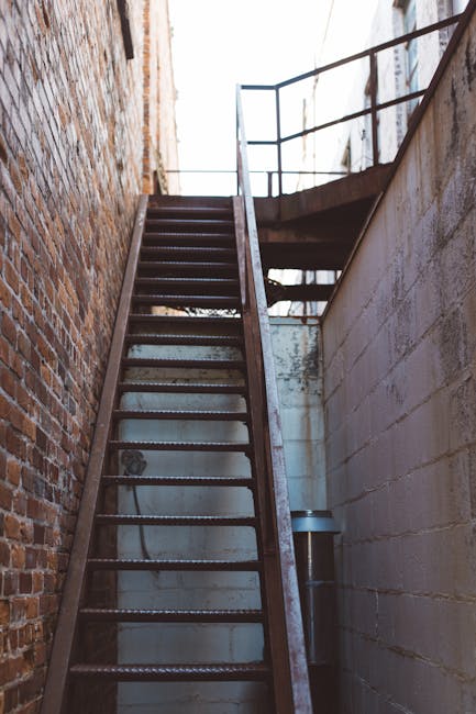A tall metal staircase with open grated steps and a railing, positioned between two brick walls, leading upward to an outdoor platform with a metal safety railing. The stairway is situated in a narrow alleyway or side passage, with the sky visible at the top. The scene is illuminated by natural daylight, highlighting the industrial appearance of the metal structure. This staircase may be part of a building's fire escape or external access route, relevant to the logistics of home relocation and moving services provided by Man With a Van Whitechapel, as seen in the context of house removals or furniture transport.