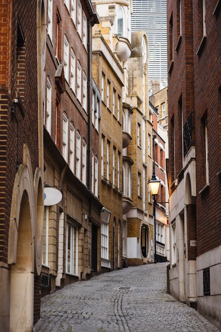 View of a narrow urban alleyway between red-brick residential buildings during daylight, with fire escape ladders and balconies made of black metal on both sides, casting shadows on the brick facades. A white van with a partially visible roof is parked at the pavement, with a person inside loading cardboard boxes and wrapped furniture using plastic and blankets. The boxes are stacked inside and near the vehicle, some on a trolley, as part of a home relocation process. The environment features clear blue sky overhead, natural lighting highlighting the textures of the brickwork and metalwork, and the scene captures a furniture transport and packing stage typical of house removals in the Whitechapel area, with Man With a Van Whitechapel providing professional removals service.