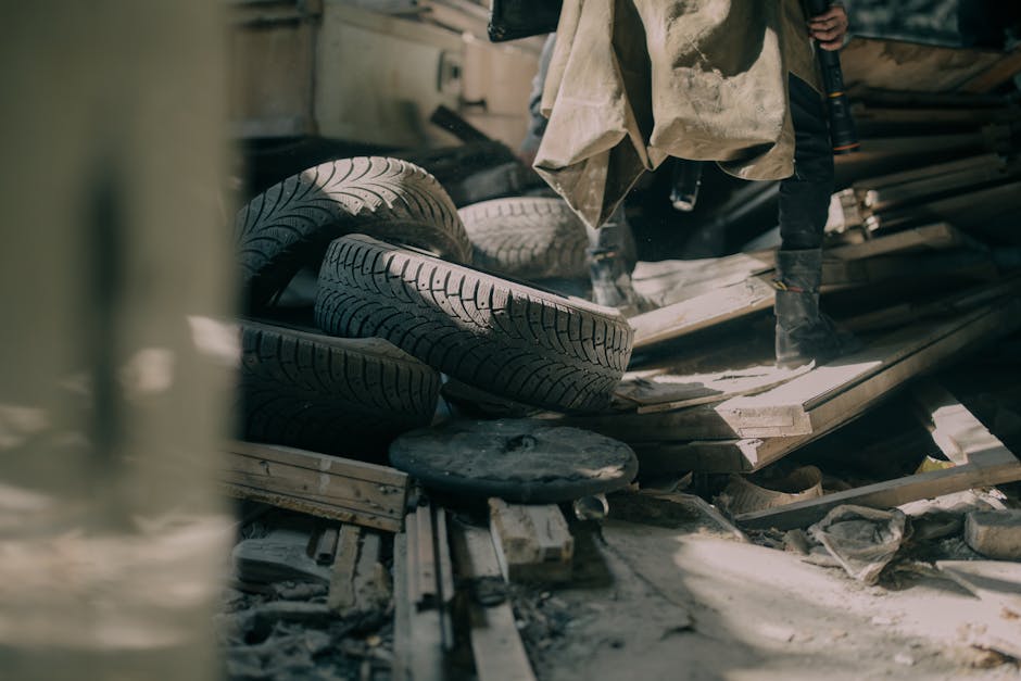 Interior of a cluttered storage area or hoarder flat showing multiple used tires stacked among various wooden planks, cardboard boxes, and scattered debris. Part of a person wearing dark clothing and black boots is visible standing on the uneven surface, possibly involved in a packing, clearing, or removal process. The environment appears dusty and disorganized, suggesting ongoing cleaning or home relocation activities. In the background, additional materials and trash are visible, with minimal lighting emphasizing the cluttered space. This scene illustrates the challenges involved in clearing hoarder flats and preparing for furniture transport or house removals, as facilitated by Man With a Van Whitechapel, specialists in removals and moving services.