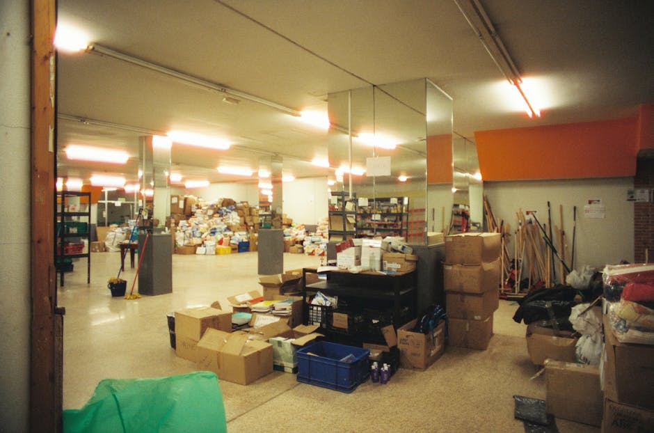 Inside a large, well-lit storage or warehouse area filled with numerous cardboard boxes of varying sizes, some stacked on top of each other, and others scattered across the floor. The space features numerous metal shelving units loaded with additional boxes and miscellaneous items. In the foreground, there are loose boxes, plastic crates, and packaging materials such as bubble wrap and foam. Several items like bags of textiles or soft materials are piled on and around the boxes. To the right, there are cleaning tools such as brooms and mops leaning against the wall. The background includes a long stretch of shelving with more packaging supplies and stored goods, creating a busy and cluttered environment typical of a house clearance or packing process during a property removal. This image depicts a scene at a property mentioned in the 'Clearing Hoarder Flats in Whitechapel' article by Man With a Van Whitechapel, illustrating the packing and loading phase of home relocations or furniture transport services.