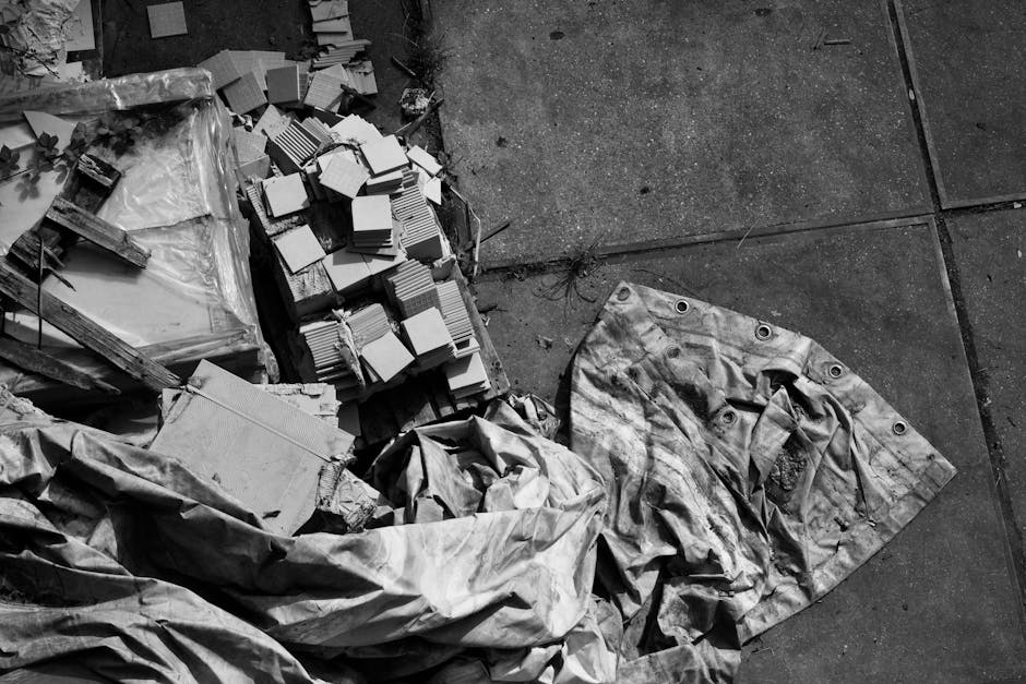 Interior of a cluttered storage area or hoarder flat showing multiple used tires stacked among various wooden planks, cardboard boxes, and scattered debris. Part of a person wearing dark clothing and black boots is visible standing on the uneven surface, possibly involved in a packing, clearing, or removal process. The environment appears dusty and disorganized, suggesting ongoing cleaning or home relocation activities. In the background, additional materials and trash are visible, with minimal lighting emphasizing the cluttered space. This scene illustrates the challenges involved in clearing hoarder flats and preparing for furniture transport or house removals, as facilitated by Man With a Van Whitechapel, specialists in removals and moving services.