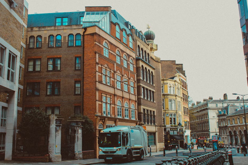 A street scene in Whitechapel featuring several multi-storey buildings with a mix of brick and ornate facades. A light blue van with the branding 'Dennis' is parked on the street, positioned close to a building entrance. Nearby, a small stack of cardboard boxes wrapped in plastic and secured with packing tape is placed on the pavement, indicating packing and moving preparations by Man With a Van Whitechapel. The street is lined with bollards and bicycles locked in racks, with street lamps and a traffic sign visible in the background. The environment appears urban, with a cloudy sky overhead, and the scene captures the loading process of furniture and boxes during a house or office relocation. Inside the entrance of one of the buildings, visible through an open doorway, are partially visible furniture pieces wrapped in protective blankets, typical in professional removals services. Overall, the image reflects an active moving day with furniture transport and logistical coordination, as part of a home or commercial move in the Whitechapel area.