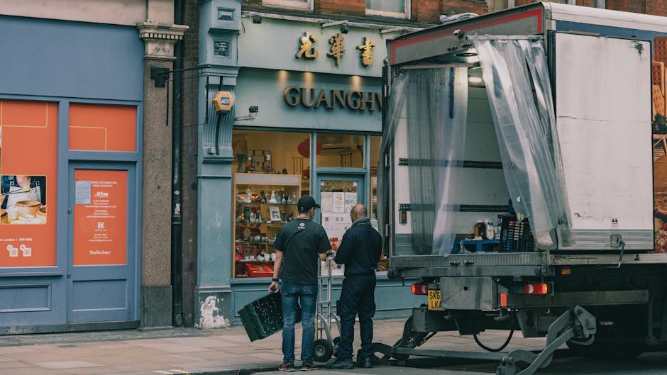 A street scene in Whitechapel featuring several multi-storey buildings with a mix of brick and ornate facades. A light blue van with the branding 'Dennis' is parked on the street, positioned close to a building entrance. Nearby, a small stack of cardboard boxes wrapped in plastic and secured with packing tape is placed on the pavement, indicating packing and moving preparations by Man With a Van Whitechapel. The street is lined with bollards and bicycles locked in racks, with street lamps and a traffic sign visible in the background. The environment appears urban, with a cloudy sky overhead, and the scene captures the loading process of furniture and boxes during a house or office relocation. Inside the entrance of one of the buildings, visible through an open doorway, are partially visible furniture pieces wrapped in protective blankets, typical in professional removals services. Overall, the image reflects an active moving day with furniture transport and logistical coordination, as part of a home or commercial move in the Whitechapel area.