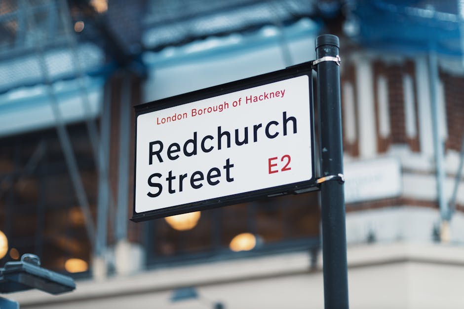 A close-up view of a street sign mounted on a black pole, displaying the name 'Redchurch Street' in bold black text, with the postcode 'E2' in red. The sign includes the additional information 'London Borough of Hackney' above the street name. The background and surroundings feature part of a building with brick and glass elements, and a blue scaffolding structure overhead, indicating an urban environment. Visible in the scene are some out-of-focus warm-lit indoor lights, suggesting nearby commercial or residential properties. The image captures a moment typical of a home relocation or moving process in the Whitechapel area, where a man with a van business might be involved in furniture transport or packing and moving activities, with the signage highlighting the specific postcode area for targeted services.