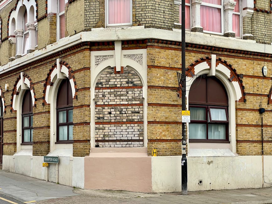 A corner view of a multi-storey Victorian-style brick building with arched windows featuring white trim and dark brown frames. The building's facade includes decorative red and white bricks and beige plaster detailing around the windows and at the corner. In front of the building, a narrow sidewalk runs parallel to the pavement, where a black lamppost with a yellow warning sign and a parking restriction sign is visible. The building is situated at the intersection of Crawford Road, indicated by a green street name sign attached to the wall near the corner. The ground level has a white skirting, and two windows on the lower floor reveal cream curtains closed behind the glass. The exterior suggests it is an older residential or mixed-use property. This setting may relate to house removals or furniture transport, as handled by Man With a Van Whitechapel, incorporated into detailed urban moving processes.