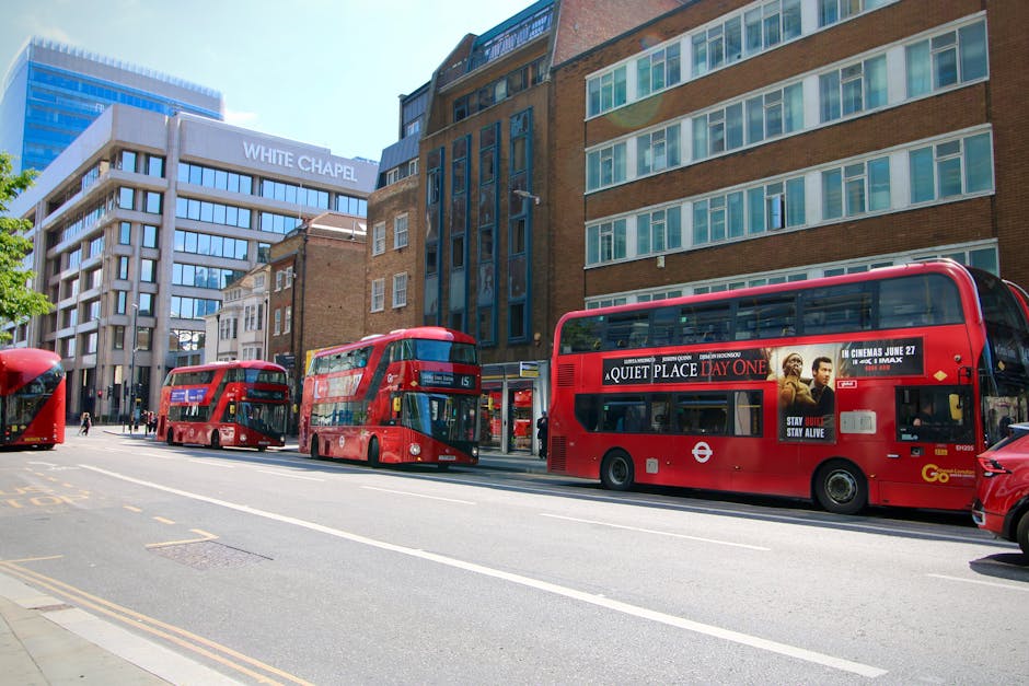 A street scene in Whitechapel showing three red double-decker buses parked along the curb, with a bus stop visible on the pavement. Behind the buses, there are multi-storey buildings, including a mode