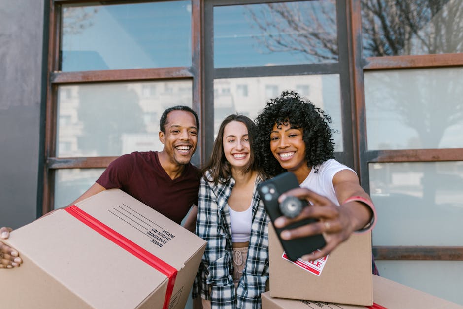 Three individuals standing outdoors in front of a building with large glass windows, smiling and taking a selfie. The person on the left, a man wearing a maroon T-shirt, holds a cardboard box sealed with red tape. The woman in the middle, with long brown hair and a plaid shirt, is smiling widely. The woman on the right, with curly black hair and a white T-shirt, holds a smartphone and is partly extending her arm to take the photo. The scene suggests a home relocation or packing process, with moving boxes visible, and the background featuring reflections of nearby buildings and trees, indicating daytime. This image, associated with Man With a Van Whitechapel, subtly relates to furniture transport and packing and moving services in the context of local removals near Whitechapel Gallery, E1.