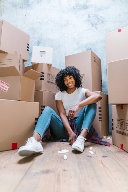 A young woman with curly black hair, wearing a white t-shirt, blue jeans, and white sneakers, is sitting on a wooden floor amidst numerous unpacked cardboard boxes of various sizes with printed labels and tape, some marked as fragile. She is smiling and relaxed, with her arms resting on her knees. The background features a textured light blue wall with signs of wear or paint splatter, and the setting appears to be a room prepared for moving or home relocation. Several packed items, including cardboard boxes and packing materials, imply an ongoing packing and moving process. The image captures the interior environment during the moving logistics phase, aligning with house removal services provided by Man With a Van Whitechapel, supporting the themes of furniture transport, packing, and home relocation in the Whitechapel area.