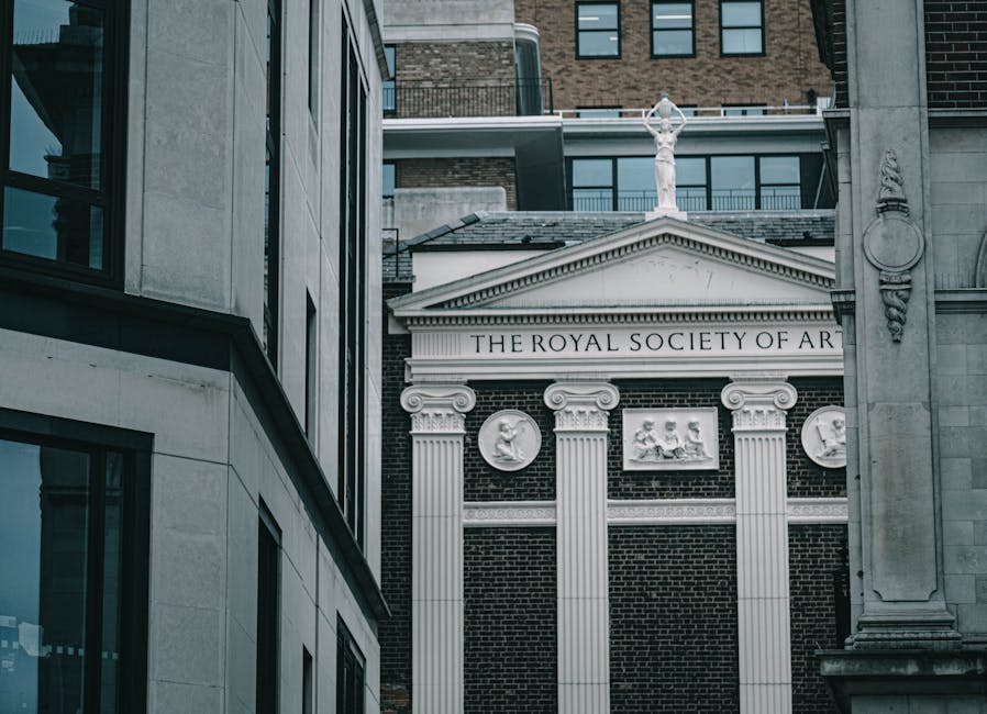 Photograph of the facade of a historic building with a brick exterior, featuring decorative white columns and medallions embedded in the wall, with the partially visible inscription 'THE ROYAL SOCIETY OF ART'. A triangular pediment is situated above the entrance, topped with a classical statue of a standing figure holding a staff. The building is flanked by other structures, including modern buildings with large windows and balconies at the rooftops, indicating an urban setting in Whitechapel. The scene is captured during daylight with natural lighting, emphasizing the architectural details. While the image emphasizes the architectural features, it subtly relates to the context of house removals and furniture transport in the area, relevant to services like those provided by Man With a Van Whitechapel for house and institutional relocations, including moving art and delicate items.