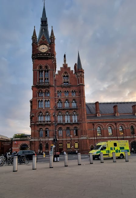 The image depicts the historic red-brick building of the Royal London Hospital with its prominent clock tower featuring multiple spires, set against a cloudy sky during late afternoon or early evening. In the foreground, a paved area with metal bollards separates the street from the hospital. A man with a white van, part of Man With a Van Whitechapel's removals fleet, is involved in the home relocation process, loading rectangular cardboard boxes and wrapped furniture items onto the vehicle. The boxes are secured with plastic wrapping or packing paper, and some are stacked on trolleys or positioned on the pavement, ready for transport. Nearby, bicycles are parked, and the yellow-green emergency vehicle is visible on the street, indicating active usage of the area. The scene captures a typical furniture transport and packing process during a professional moving service in the Whitechapel area, supporting the logistics of same-day removals for residential relocations related to the PAGE_TITLE.