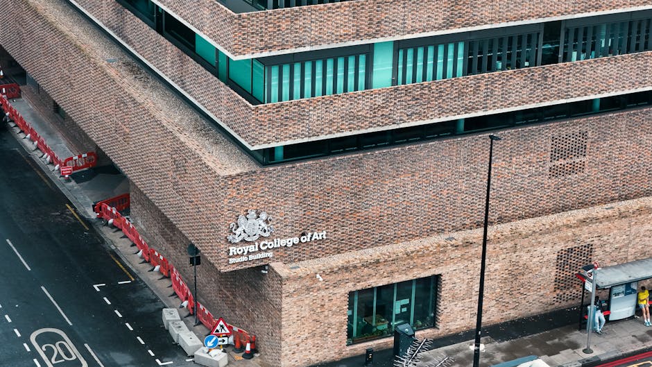 Photograph of the facade of a historic building with a brick exterior, featuring decorative white columns and medallions embedded in the wall, with the partially visible inscription 'THE ROYAL SOCIETY OF ART'. A triangular pediment is situated above the entrance, topped with a classical statue of a standing figure holding a staff. The building is flanked by other structures, including modern buildings with large windows and balconies at the rooftops, indicating an urban setting in Whitechapel. The scene is captured during daylight with natural lighting, emphasizing the architectural details. While the image emphasizes the architectural features, it subtly relates to the context of house removals and furniture transport in the area, relevant to services like those provided by Man With a Van Whitechapel for house and institutional relocations, including moving art and delicate items.