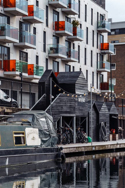 A modern multi-storey residential building with white and grey facades, featuring balconies with glass railings and orange accent panels, situated alongside a canal. In the foreground, a black boat is docked, partially covered with a grey tarp. Adjacent to the water, there is a small black wooden structure with a sign reading 'WHARF' and a string of outdoor lights hanging above. Bicycles are parked underneath the structure, along with some coiled hoses and containers. The setting appears to be part of a home relocation or moving process by [COMPANY_NAME], with the scene capturing the loading area near the water's edge, highlighting the transport and packing environment involved in furniture transport and house removals within the urban waterfront location.