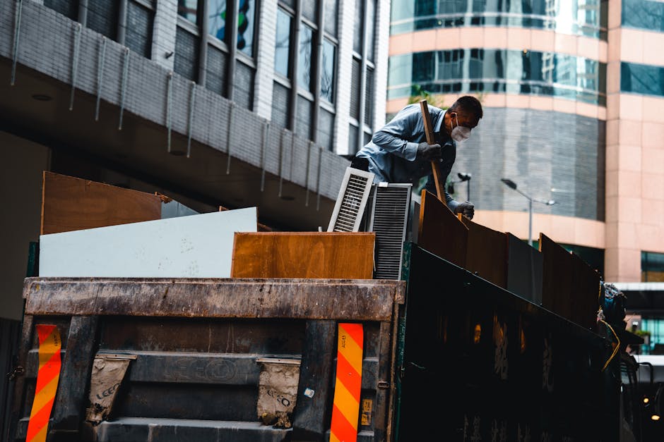 A man wearing a face mask and gloves is standing on the back of a large, open-top black truck with orange and yellow reflective markings, in an urban setting with modern glass and brick buildings in the background. He is lifting or adjusting furniture and packed items, which include wooden cabinets, vent units, and cardboard boxes, some wrapped in plastic film or blankets for protection. The truck is positioned on a paved area near a building entrance, with various household items ready for removal or transportation. Nearby, there are additional packing materials such as cardboard and plastic wrapping, indicating a home relocation or furniture transport operation. The scene captures the loading process as part of professional removals, carried out by [COMPANY_NAME], demonstrating their expertise in logistics, packing, and moving furniture efficiently in compliance with local council rules, relevant to services like skip hire versus licensed removals in Whitechapel.