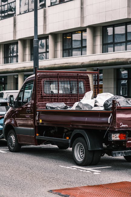 A dark red delivery van with an open flatbed parked on a city street in Whitechapel, with a modern office building in the background. The flatbed is loaded with various packing materials, including large plastic bags, boxes wrapped in protective covering, and some furniture items secured with straps. The van belongs to Man With a Van Whitechapel, a professional removal services company, and is positioned near a curb with a pavement visible in the foreground. The scene reflects the logistics involved in home relocation, with items prepared for transportation and loading processes typical in furniture transport or packing and moving activities. Nearby, a black pole is mounted on the pavement, and other parked vehicles can be seen in the vicinity, emphasizing an urban environment suitable for house removals or specialist relocation services.