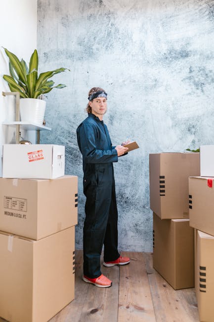 A man wearing a navy-blue coverall and a headband is standing inside a room with a textured, light blue wall behind him. He is holding a clipboard and pen, appearing to take inventory or note details. Surrounding him are several cardboard boxes of various sizes, some labeled with text such as 'glass,' and others plain; these are stacked on the wooden floor. To his left is a white shelf with a potted plant featuring large green leaves. The scene suggests a home relocation or packing process, with the man preparing for a furniture transport or moving service. The environment is well-lit, and the boxes are ready for loading into a van, reflecting the logistics involved in house removals, managed by services such as Man With a Van Whitechapel.