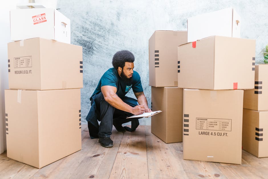 A man with an afro hairstyle is kneeling on a wooden floor, using a clipboard and pen to take inventory of a large collection of unpacked cardboard boxes of various sizes inside a property. The boxes are sealed with tape and have printed labels indicating contents such as bedroom items and furniture, with some stacked on top of each other while others are directly on the floor. The room has a textured light blue wall, and natural light illuminates the scene. This image depicts the packing and home relocation process, illustrating furniture transport and moving logistics. The presence of boxes and the man's documentation activity highlights aspects of packing and moving services offered by Man With a Van Whitechapel, aligned with house removals and permit requirements for relocation in Tower Hamlets.