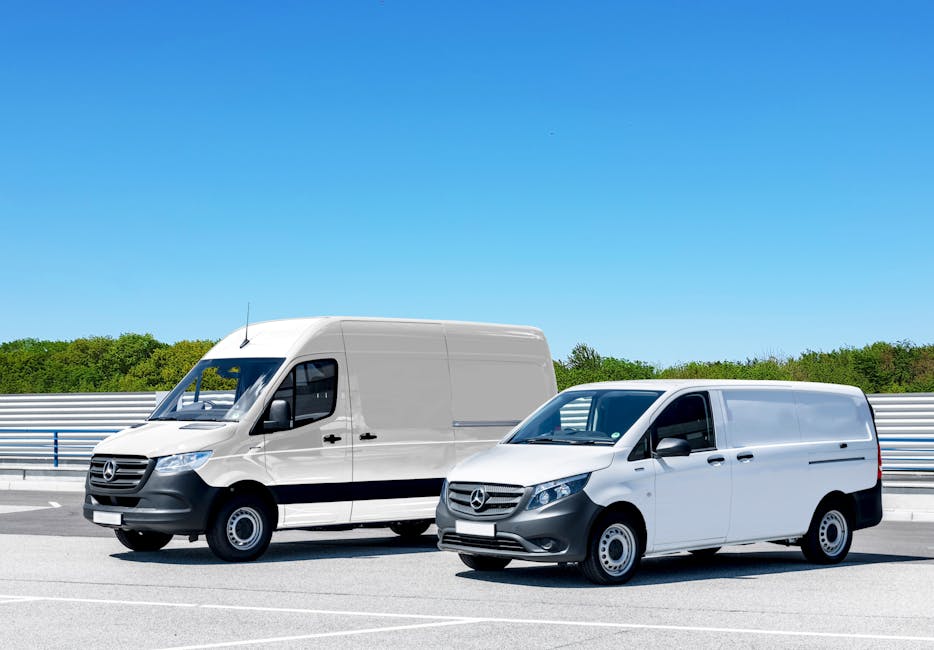 Two white Mercedes-Benz vans parked side by side on a paved road during daytime, with a clear blue sky and green trees in the background. The larger van on the left is positioned partly inside a prope