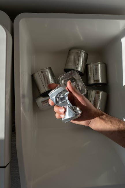 A person's hand holding a crumpled aluminium beverage can above an open white freezer drawer, with several more empty cans placed inside the freezer. The scene is set outdoors or in a well-lit area, potentially during a home relocation process involving packing or disposal of recyclable materials. The image depicts a typical step in preparing household items, such as empty cans, for removal or recycling as part of house moving or furniture transport services offered by Man With a Van Whitechapel, with the focus on handling recyclable packaging during packing and moving logistics.