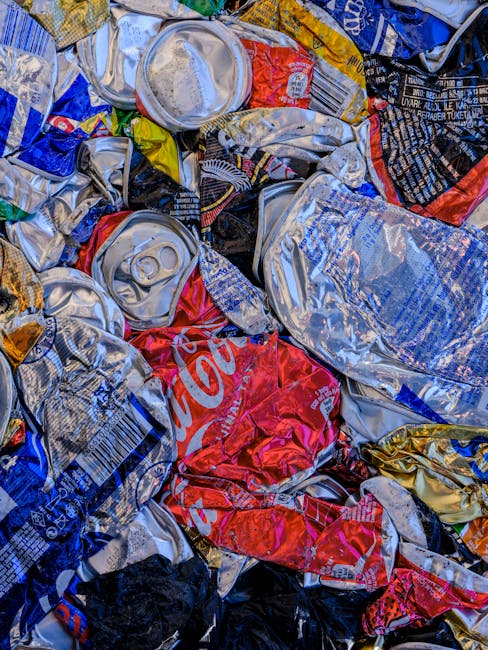 Close-up view of a large pile of crumpled aluminum cans and crushed beverage containers, including soda cans with visible pull tabs, in various colours such as silver, red, blue, and gold. The cans are mixed with some partially flattened and others intact, showing labels and branding, including a prominent red Coca-Cola can. The cans are resting on a surface that appears to be inside a property or near a waste collection area, with some edges of packaging or plastic wrap visible. The image captures the cluttered appearance typical of recyclables prepared for disposal or collection, highlighting the importance of proper waste sorting during a house removal or relocation process. This scene refers to recycling guidelines relevant to house removals in Whitechapel, as indicated by the webpage information from Man With a Van Whitechapel.